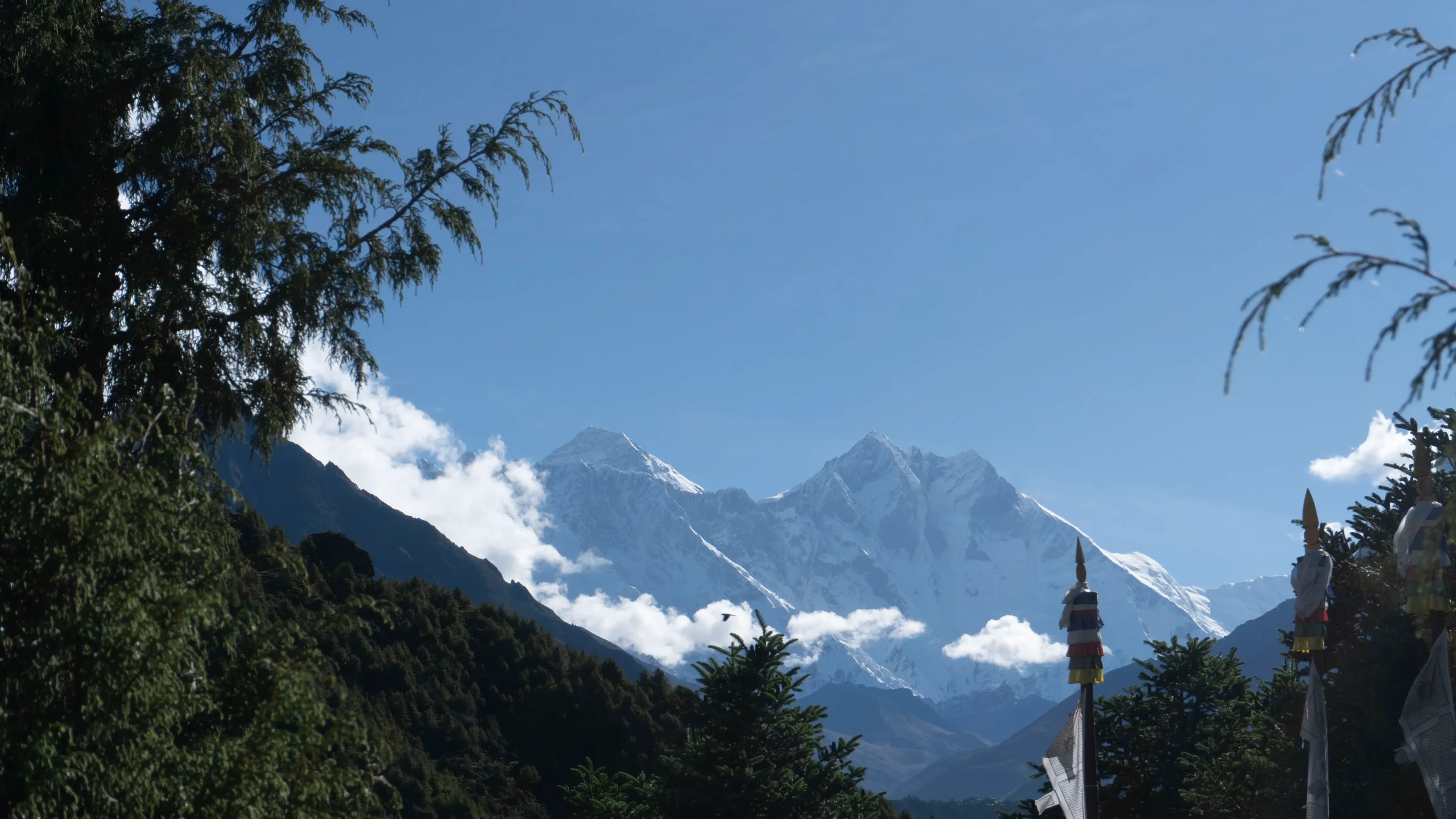 Snow-covered peaks of Mount Everest and Lhotse rising behind green forested hills and Buddhist prayer flags in Nepal under a clear blue sky.
